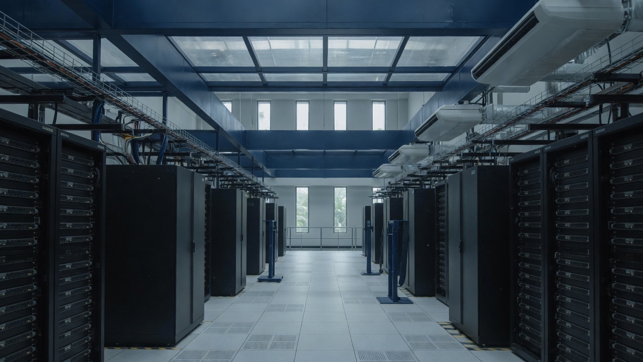 Data hall aisle between two rows of server racks, with overhead cable trays and daylight at the far windows